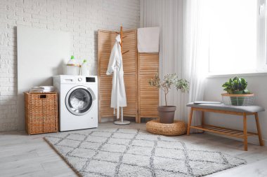 Çamaşır makinesi, bornoz ve hasır sepeti olan modern çamaşır odasının içinde.