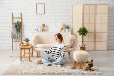 Evde dizüstü bilgisayar kullanan sevimli köpekli genç bir kadın.