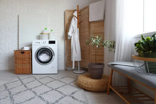 Interior of modern laundry room with washing machine, bathrobe on rack and wicker basket
