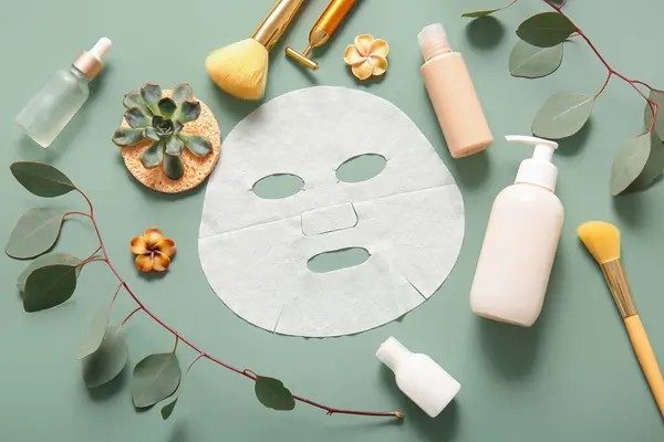 Facial sheet mask with different cosmetic products and leaves on green background