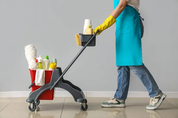 Female janitor with trolley of cleaning supplies near light wall