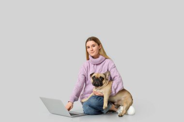 Gri arka planda oturan dizüstü bilgisayarı ve sevimli köpeğiyle güzel genç bir kadın.