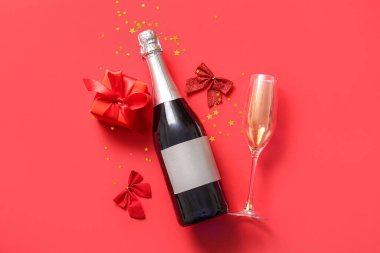 Kırmızı arka planda cam, fiyonk ve Noel hediye kutularıyla dolu bir şişe şampanya.