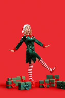 Kırmızı arka planda Noel hediyeleri olan elf gibi giyinmiş mutlu genç bir kadın.