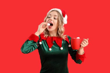 Elf gibi giyinmiş genç bir kadın elinde bir fincan sıcak çikolata ve kırmızı arka planda marşmelov tutuyor.