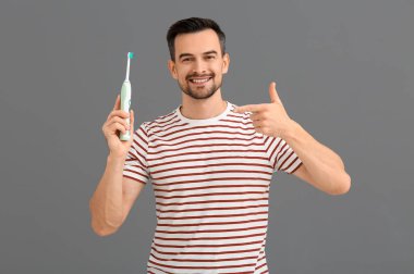 Gri arkaplanda elektrikli diş fırçasını gösteren yakışıklı bir adam.