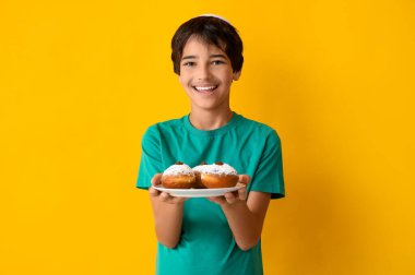 Kipa 'lı küçük çocuk elinde lezzetli donutlarla sarı arka planda bekliyor. Hanuka kutlaması