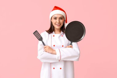 Noel Baba şapkalı güzel kadın aşçı. Pembe arka planda kızartma tavası ve spatulası var.