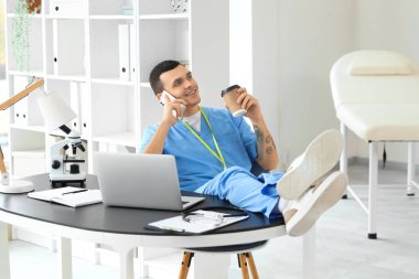 Klinikte elinde kahve fincanı olan bir erkek doktor cep telefonuyla konuşuyor.
