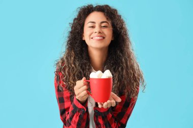 Güzel, mutlu, Afro-Amerikan bir kadın. Mavi arka planda marşmelov kaplı bir fincan kakao tutuyor.