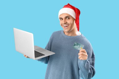 Mavi arka planda dizüstü bilgisayarı ve kredi kartı olan Noel Baba şapkalı yakışıklı bir genç. Noel alışveriş konsepti
