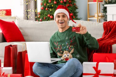 Noel için dekore edilmiş odada hediye kartı, hediye kutuları ve laptop alışverişi yapan genç ve mutlu bir adam.