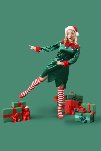 Happy young woman dressed as elf with Christmas gifts pointing at something on green background