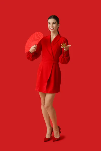 Beautiful young happy woman with fan and golden dragon figurine on red background. Chinese New Year celebration