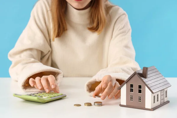 Young woman with calculator, coins and house model at table on blue background, closeup. Price rise concept