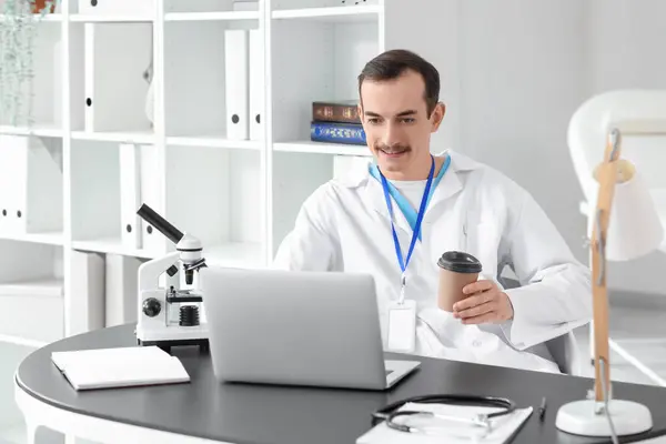 Male doctor with coffee cup working at desk in clinic