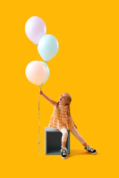 Cute little girl with beautiful balloons sitting on cube against yellow background