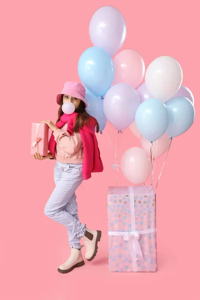 Trendy girl with gift boxes and balloons blowing bubble gum on pink background