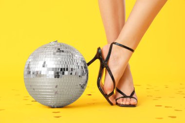 Şık topuklu kadın bacakları, disko topu ve sarı arka planda konfeti.