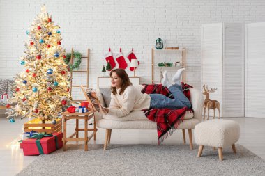 Güzel, genç, mutlu bir kadın Noel süsü verilmiş bir odada kanepede uzanıyor ve dergi okuyor.