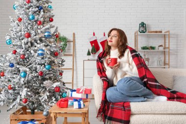 Bir fincan sıcak kakaolu güzel bir kadın Noel için dekore edilmiş bir odada koltukta oturuyor.
