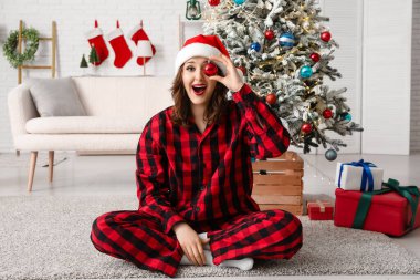 Kareli kırmızı pijamalı, Noel Baba şapkalı ve Noel balosuyla süslü evinde oturan genç ve güzel bir kadın.