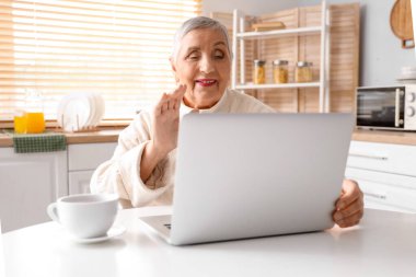 Dizüstü bilgisayarı olan yaşlı bir kadın mutfaktaki masada sohbet ediyor.