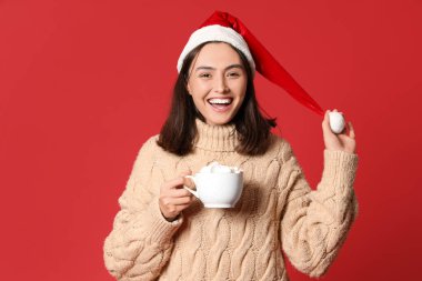 Kırmızı arka planda kakaolu Noel Baba şapkalı mutlu genç kadın.