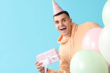 Parti şapkalı genç adam. Hediye kutusu ve mavi arka planda hava balonları.