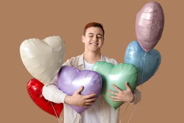 Kahverengi arka planda kalp şeklinde balonları olan mutlu genç bir adam.