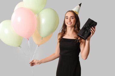 Parti şapkalı, hediye kutusu ve hava balonlarıyla mutlu genç bir kadın gri arka planda doğum gününü kutluyor.