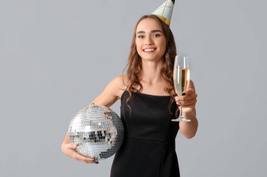 Parti şapkalı güzel bir kadın, disko topu ve bir bardak şampanyayla gri arka planda doğum gününü kutluyor.