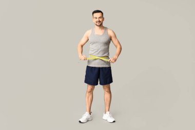 Gri arka planda belini ölçen yakışıklı, genç bir adam. Kilo verme konsepti