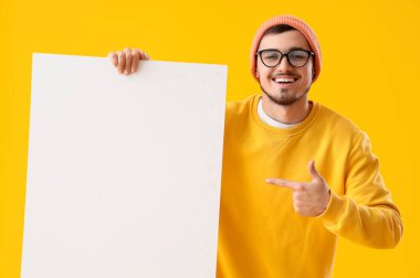 Gözlüklü genç adam sarı arka plandaki boş posterlere bakıyor.