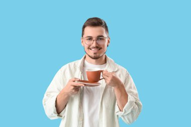 Gözlüklü genç adam mavi arka planda kahve içiyor.