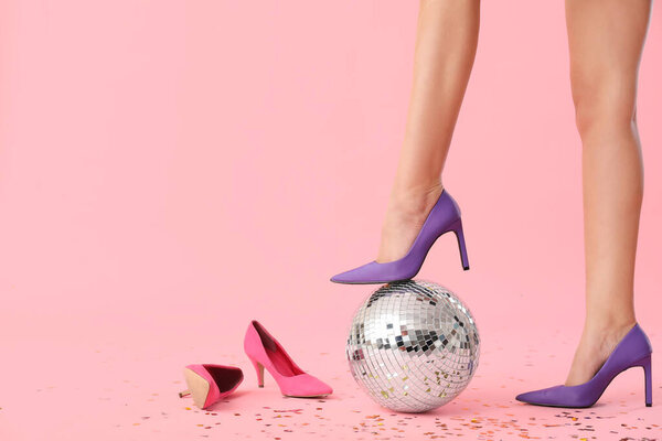 Female legs in stylish high heels with disco ball and confetti on pink background