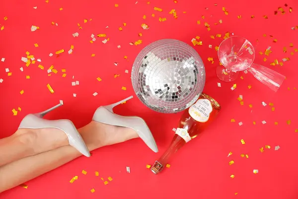 Şık topuklu kadın bacakları, disko topu ve kırmızı arka planda şampanya şişesi.