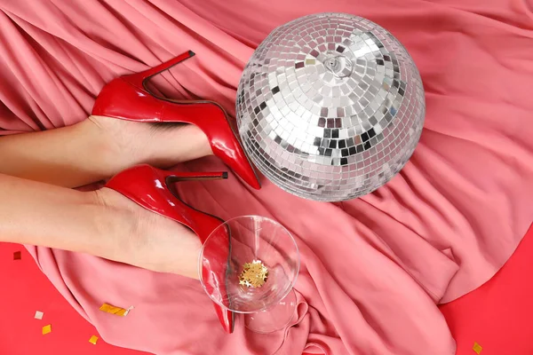 Şık topuklu kadın bacakları, disko topu ve kırmızı arka planda şampanya bardağı.