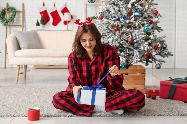 Kareli kırmızı pijamalı, geyik boynuzlu güzel genç bir kadın. Süslü evde bir fincan sıcak kakao ve Noel hediyesi.