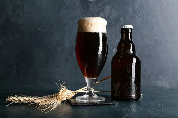 Mug and bottle of cold dark beer with wheat on table against dark grunge background