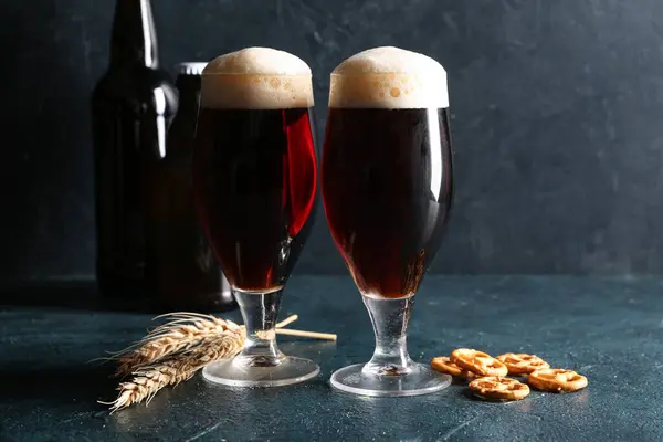 Glasses of cold dark beer with wheat and pretzels on table against dark grunge background