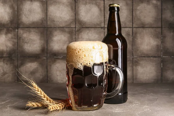 Mug and bottle of cold dark beer with wheat on table against grey tile background