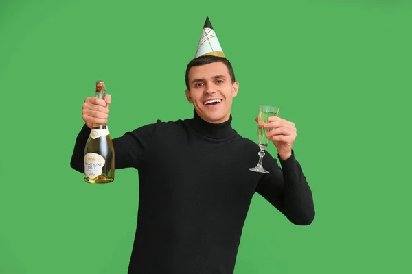 Happy young man in party hat with glass and bottle of champagne celebrating Christmas on green background