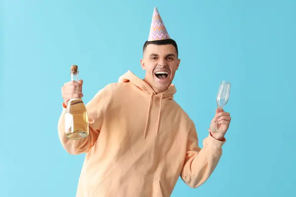 Young man in party hat with glass and bottle of champagne on blue background