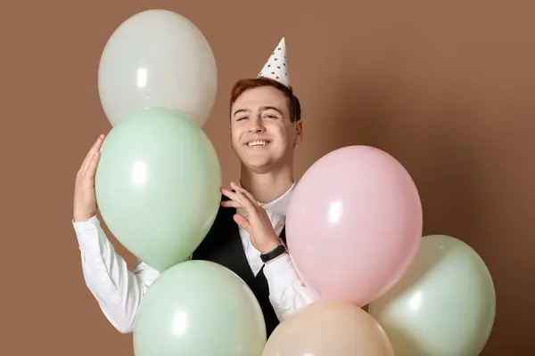 Happy young man in party hat with air balloons celebrating Birthday on brown background
