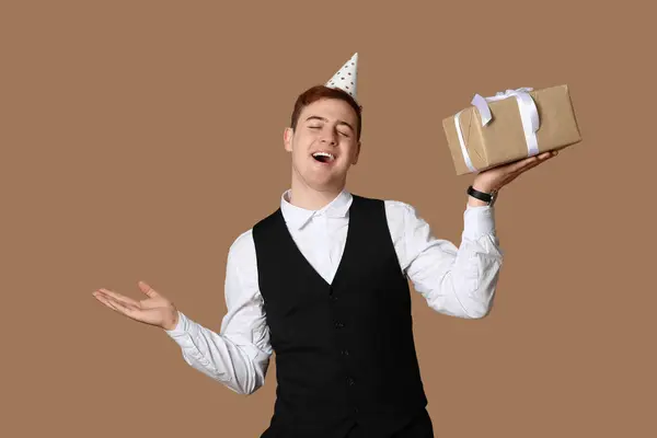 Happy young man in party hat with gift box celebrating Birthday on brown background
