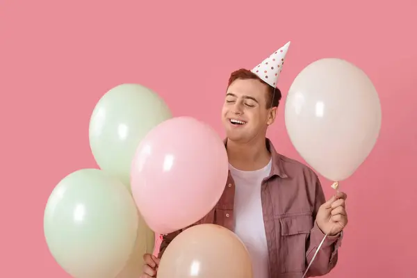 Young man in party hat with air balloons celebrating Birthday on pink background