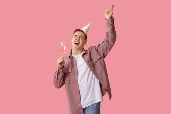 Young man with party whistle and glass of champagne celebrating Birthday on pink background