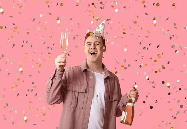 Happy young man in party hat with glass and bottle of champagne celebrating Birthday on pink background