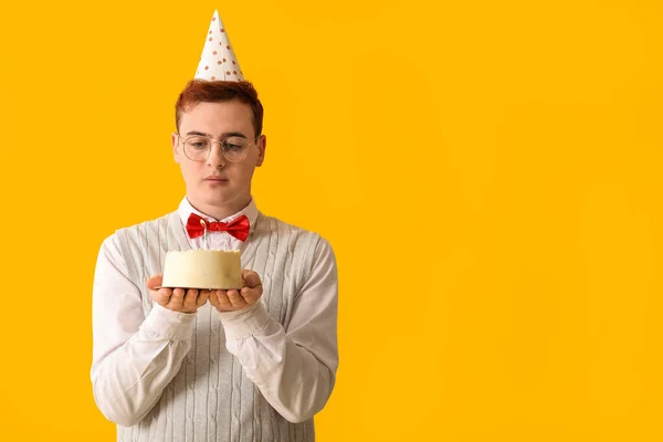 Young man in party hat with sweet cake celebrating Birthday on yellow background